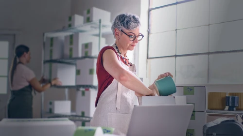 Two people working in a bright pottery studio with white shelving, one wearing a