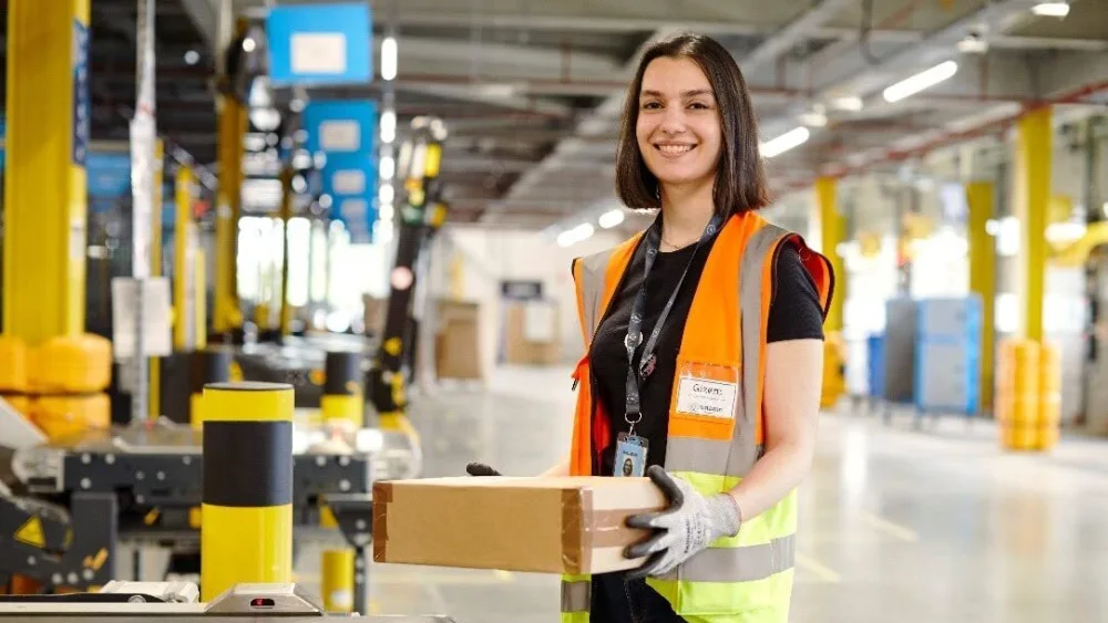 Trabajadora de almacén con chaleco de seguridad de alta visibilidad manipulando paquetes en un centro logístico de Amazon, rodeada de equipo industrial