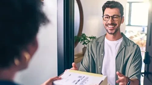 Persona con ropa casual y gafas aceptando una entrega en la puerta, intercambiando sonrisas con el repartidor