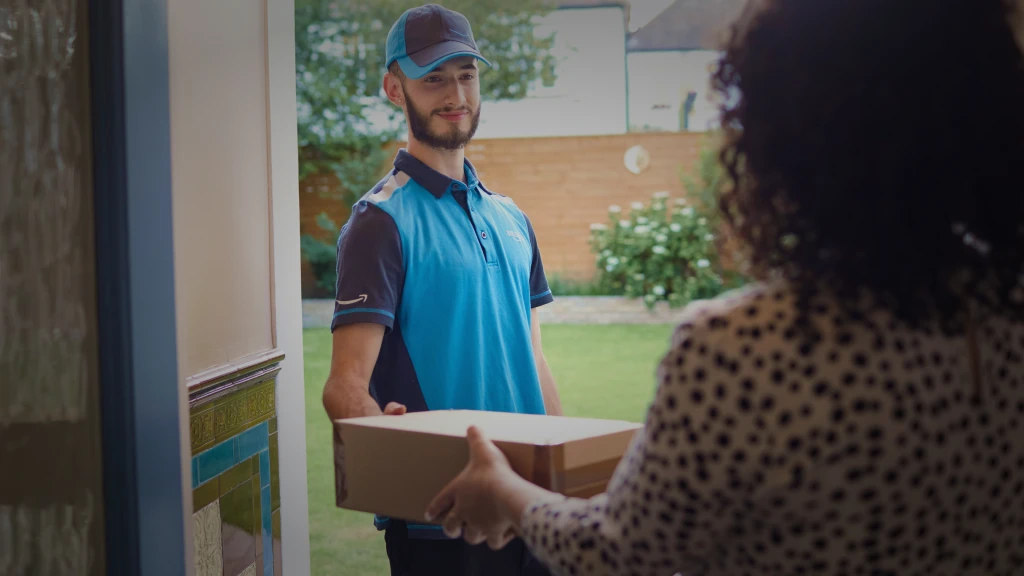 A man in a blue Amazon delivery uniform hands a package to a woman