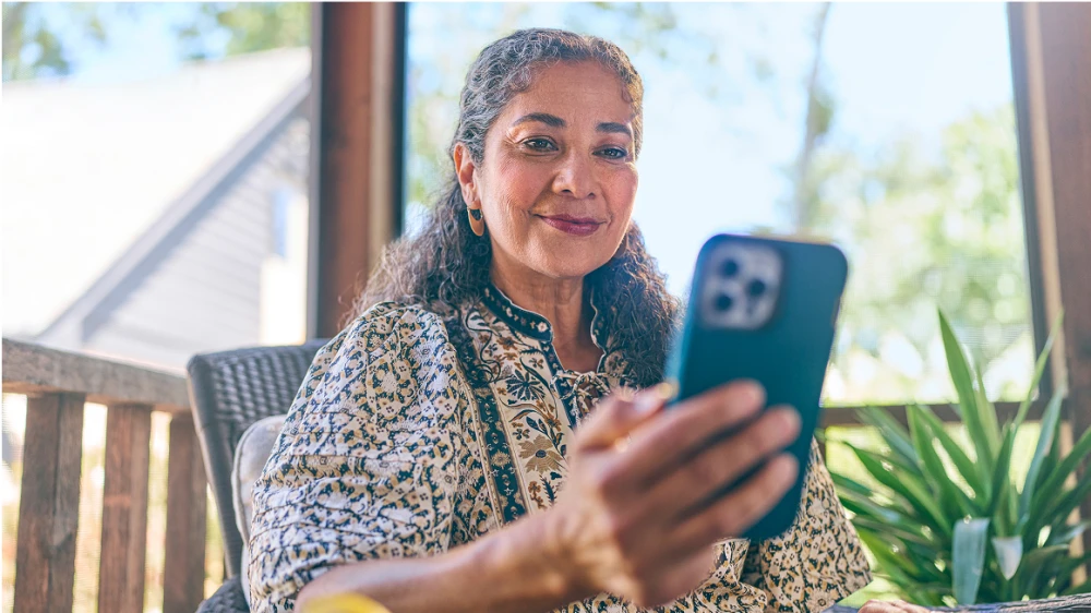 Person with curly gray hair in floral print shirt sits on porch holding smartphone, with plants and natural lighting in background