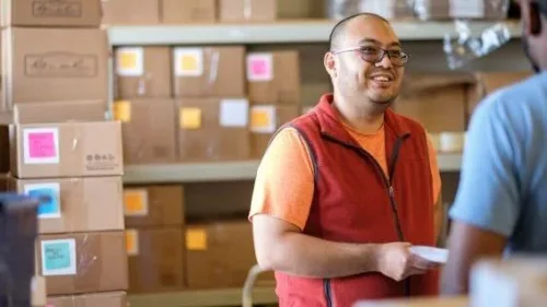 A warehouse worker in an orange shirt and red vest smiles while holding paperwork, with stacked boxes and colorful labels in background.