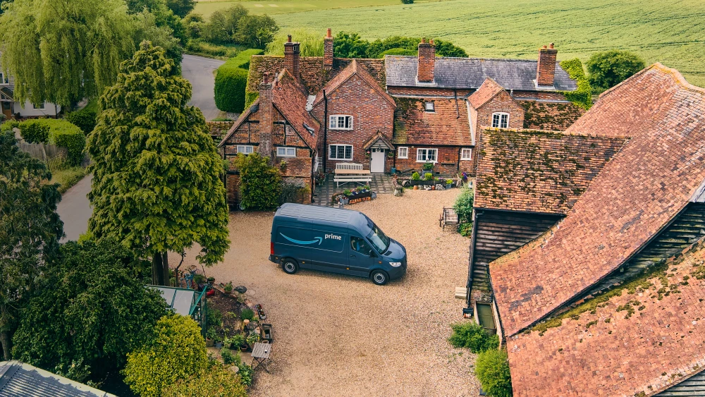 Aerial view of traditional English countryside home with brick buildings, tiled roofs, and Amazon delivery van in gravel courtyard.