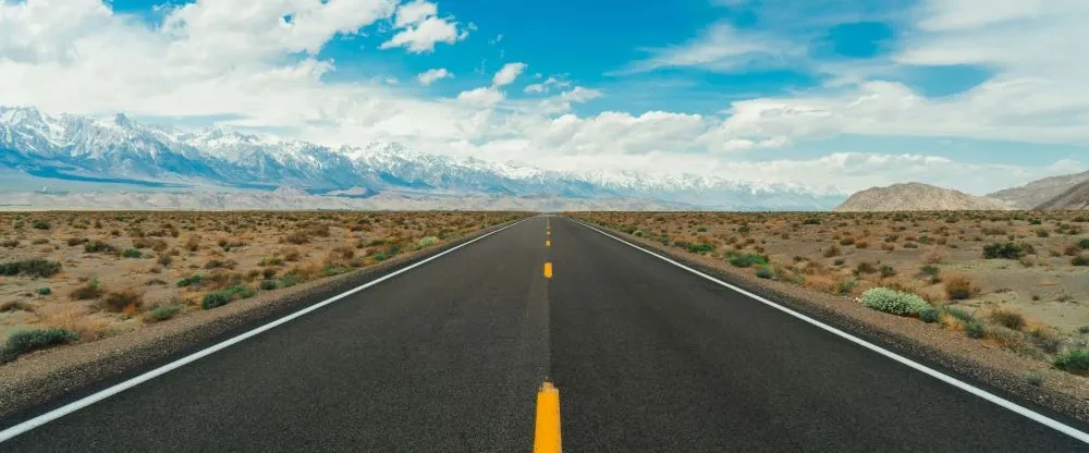 an empty road in the foreground, with patches of grass and sand on the sides. In the background, there are mountains and a clear sky. To illustrate an article on freight lanes