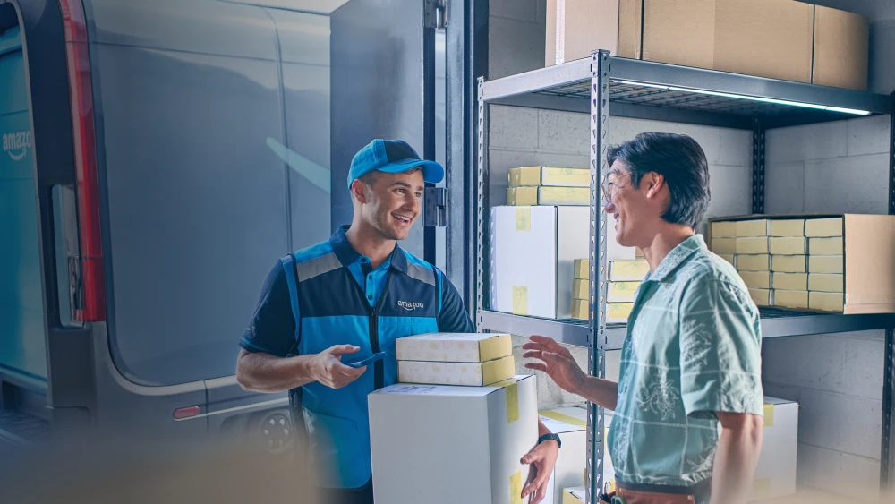 Amazon delivery driver and customer share a friendly interaction while exchanging packages in a warehouse-style setting with shelves of boxes