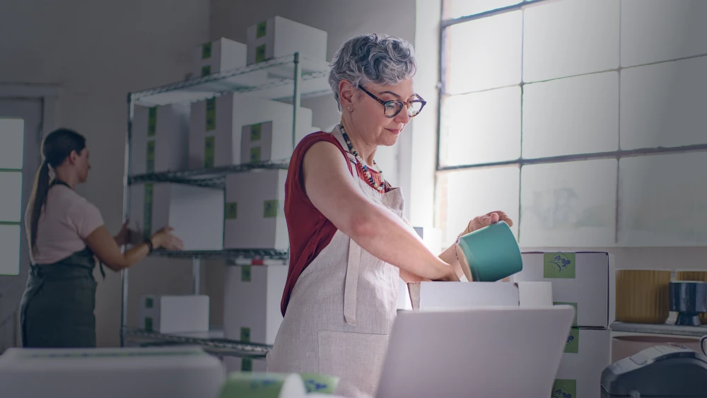 Two people working in a bright pottery studio with white shelving, one wearing an apron and packing a turquoise ceramic piece into a box.