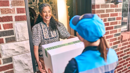 Delivery worker in bright blue uniform hands package to smiling resident at brick home's doorway