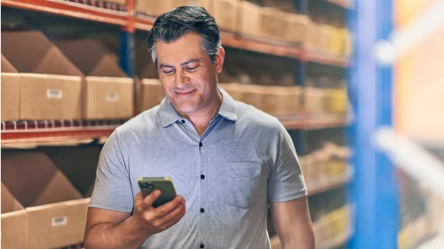 Warehouse worker in gray polo shirt checking mobile phone, standing in front of shelves stocked with cardboard boxes