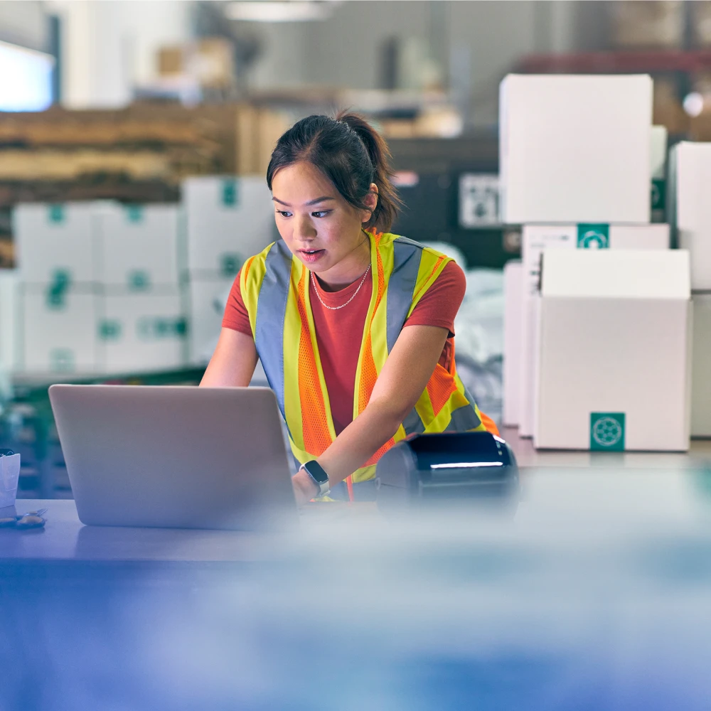 Warehouse worker in safety vest working on laptop among stacked boxes.