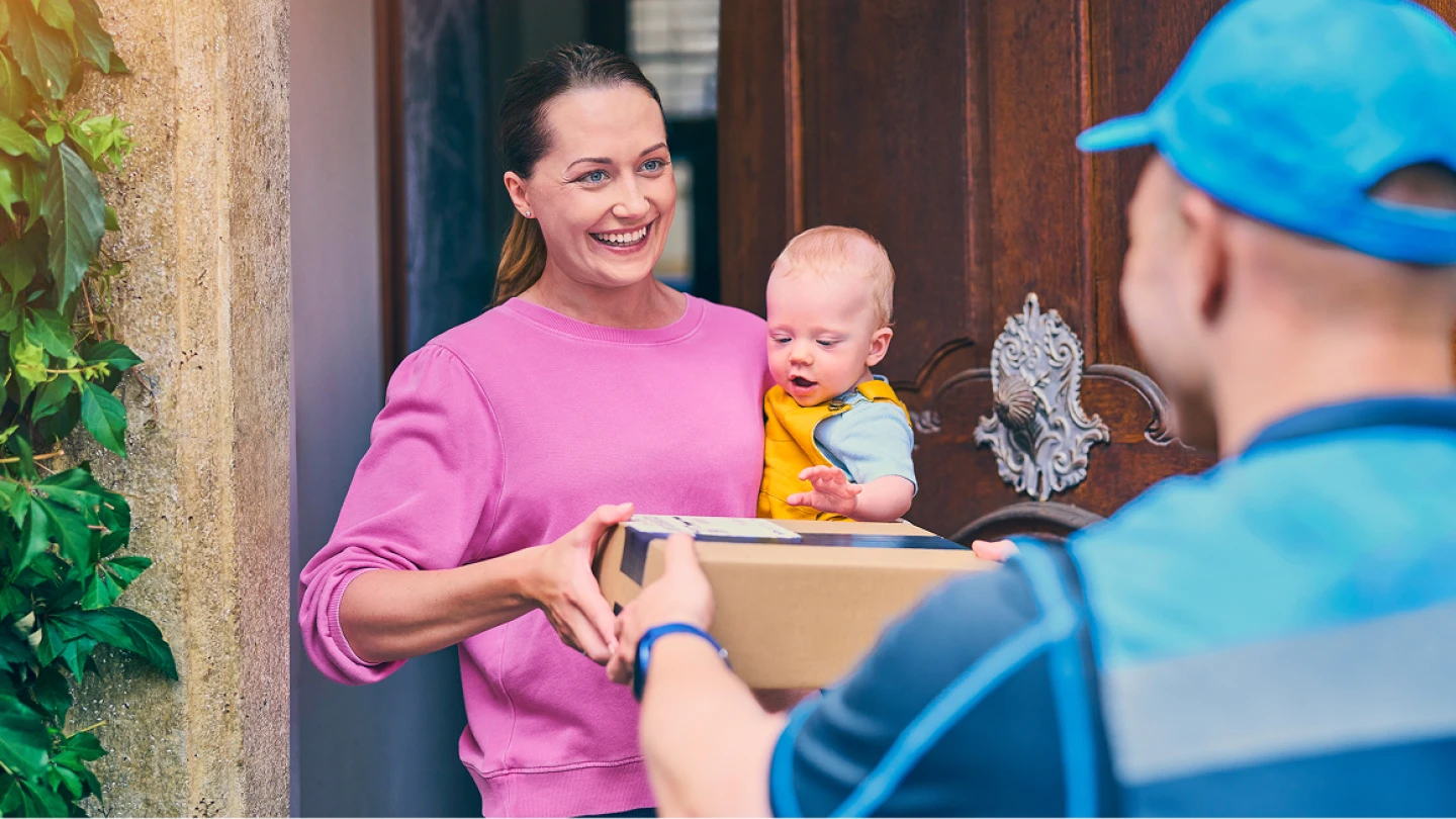 Delivery person hands package to person in pink sweater holding baby in yellow at front door of home