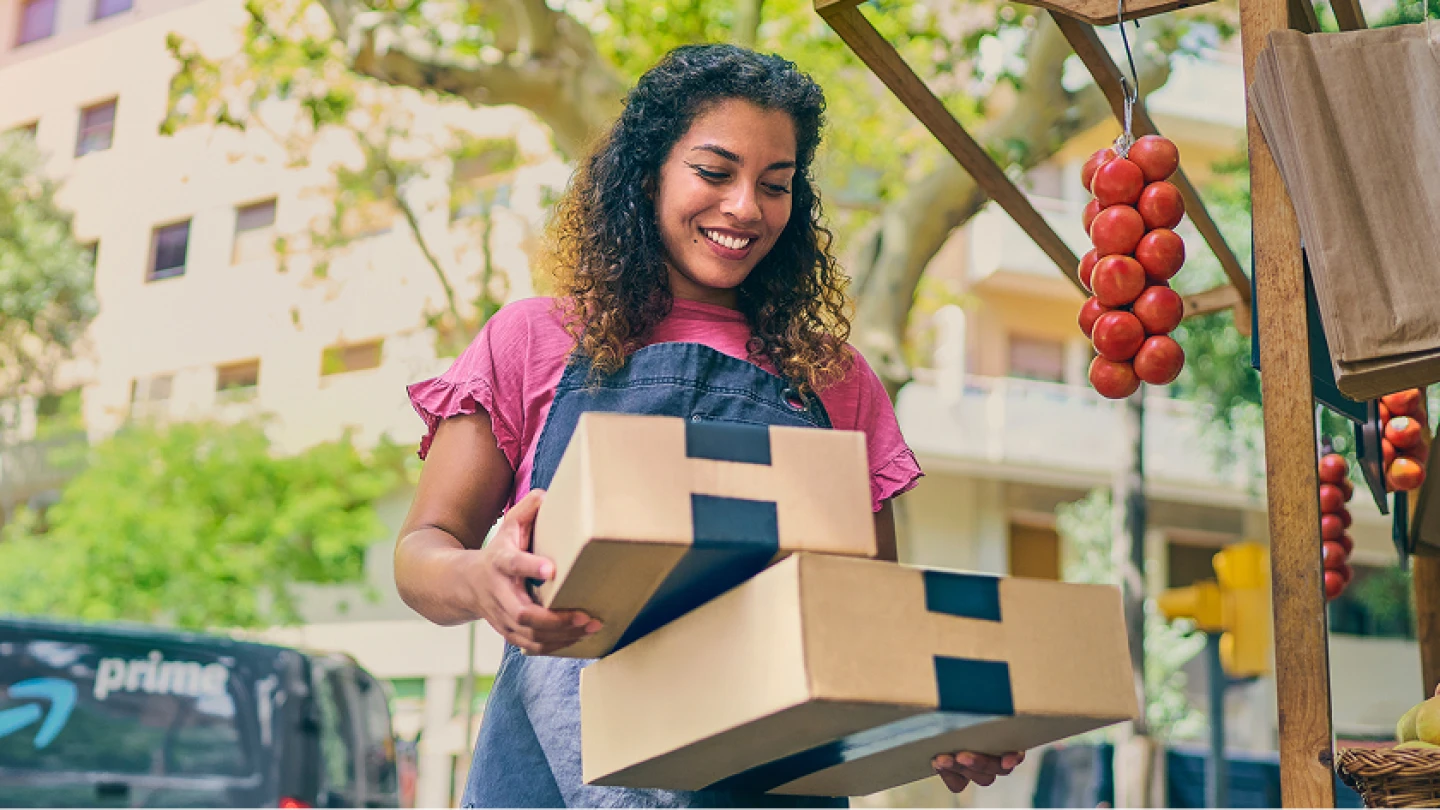 Delivery worker in denim apron carries stack of packages at outdoor market stall with hanging tomatoes and Prime van visible