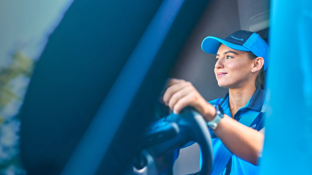 Delivery worker in blue uniform and cap smiling while sitting in vehicle, viewed through window in profile angle