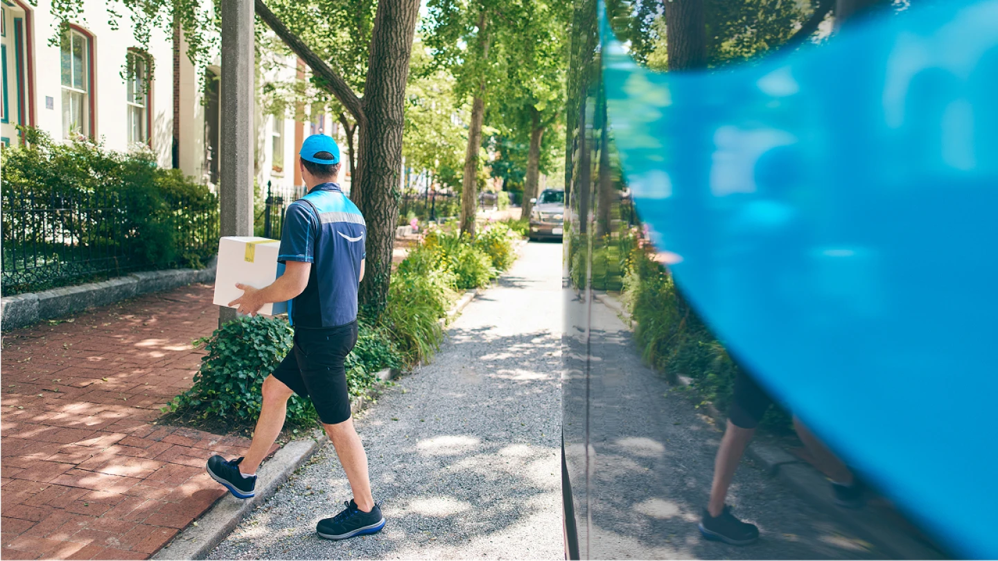 Delivery worker in blue uniform carrying package walks along tree-lined residential street with brick sidewalk on sunny day