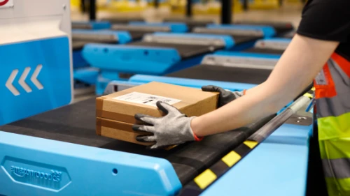 Trabajador con chaleco de seguridad coloca paquete de Amazon en sistema de cinta transportadora azul en una instalación de clasificación del almacén