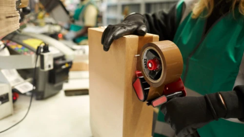 Worker in green apron and black gloves using handheld packing tape dispenser on cardboard box in warehouse setting