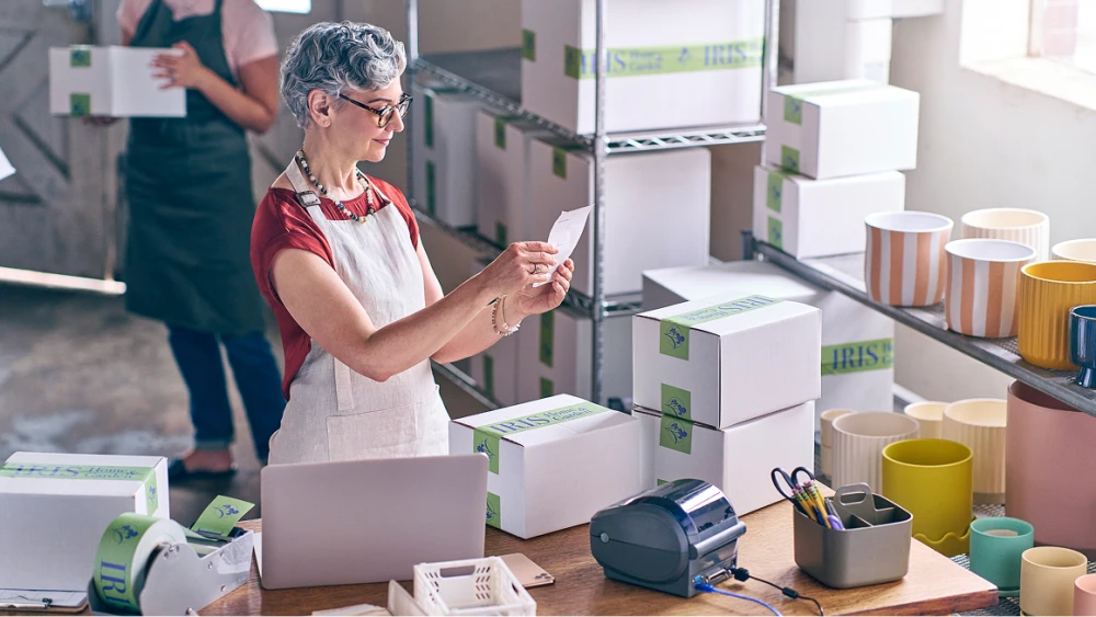 Small business owner in apron processing orders with laptop and shipping boxes on shelves
