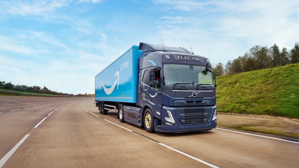 Electric Volvo semi truck with Amazon Prime blue trailer driving on highway, photographed at an angle against green hillside and blue sky