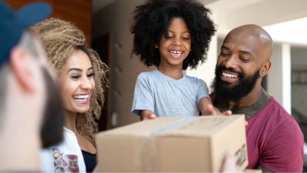 A happy family receives a delivery package at their front door, all smiling as they accept the cardboard box from someone.