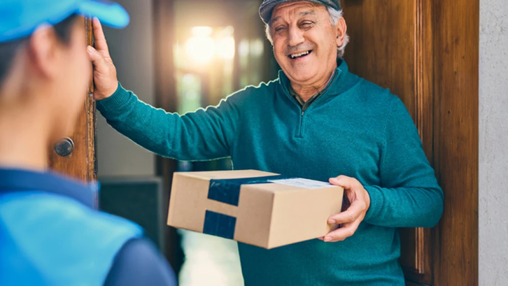 Smiling senior in green sweater receives package delivery from courier at doorway, warm interaction during home delivery service.
