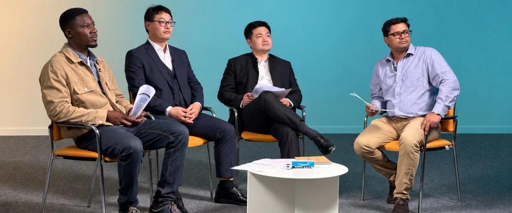 Four men seated on chairs in a panel discussion setup, holding papers, with a white table in front against a teal wall.