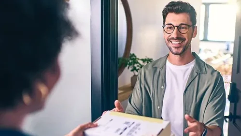 Persona con ropa casual y gafas aceptando una entrega en la puerta, intercambiando sonrisas con el repartidor