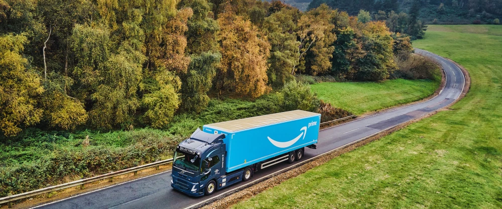 Amazon Prime delivery truck driving on curved rural road surrounded by autumn trees and green fields from aerial view.