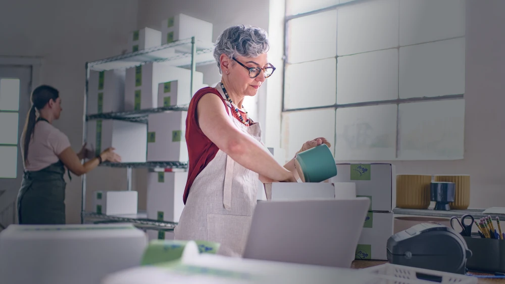 Two people working in a bright pottery studio with white shelving, one wearing an apron and packing a turquoise ceramic piece into a box.