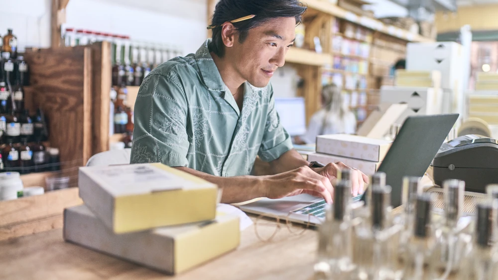 Small business owner in teal shirt works at wooden counter with laptop, boxes, and glass bottles on display in shop interior