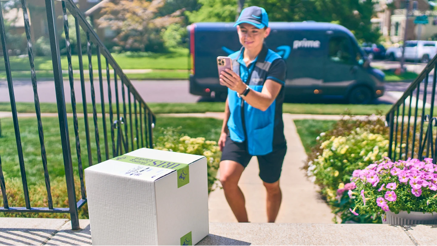 Amazon Prime delivery driver takes photo of package on her phone, with delivery van and flowers visible in background.