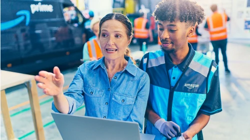 Two warehouse workers in denim and Amazon uniforms collaborate while looking at a laptop in a distribution center environment.