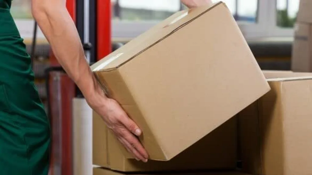 Trabajador de reparto con uniforme verde levantando y apilando cajas de cartón marrón en un almacén o centro de almacenamiento.