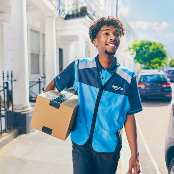 Amazon delivery worker in blue uniform vest carrying a package on a sunny residential street