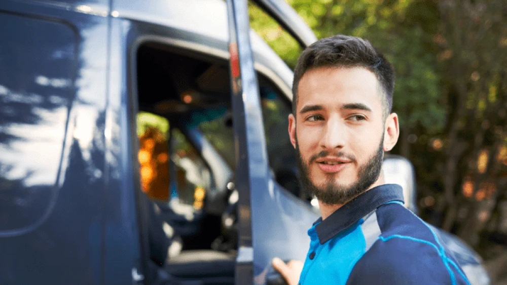 Chauffeur-livreur en uniforme bleu debout à côté d'une camionnette grise, avec des arbres visibles en arrière-plan