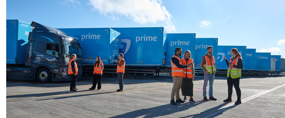 Workers in safety vests stand near Amazon Prime delivery trucks at a logistics facility under blue sky.