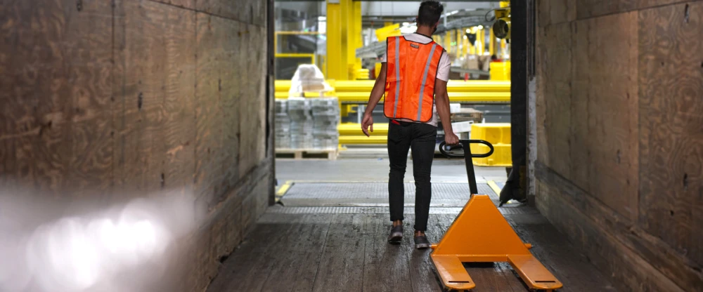 Worker in orange safety vest pulling pallet jack through industrial corridor with yellow barriers visible ahead.