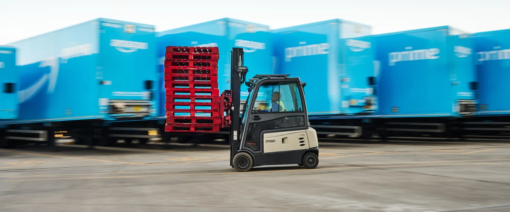 Crown forklift carrying red pallets in front of blue shipping containers at an industrial facility.