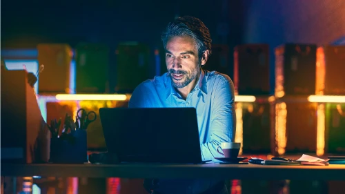 Person working late at desk with laptop, illuminated by colorful ambient lighting and screen glow in dark office setting