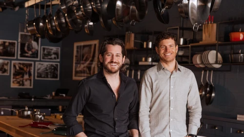 Two men, the founders of Made In, lean on a long table with cookware displayed o