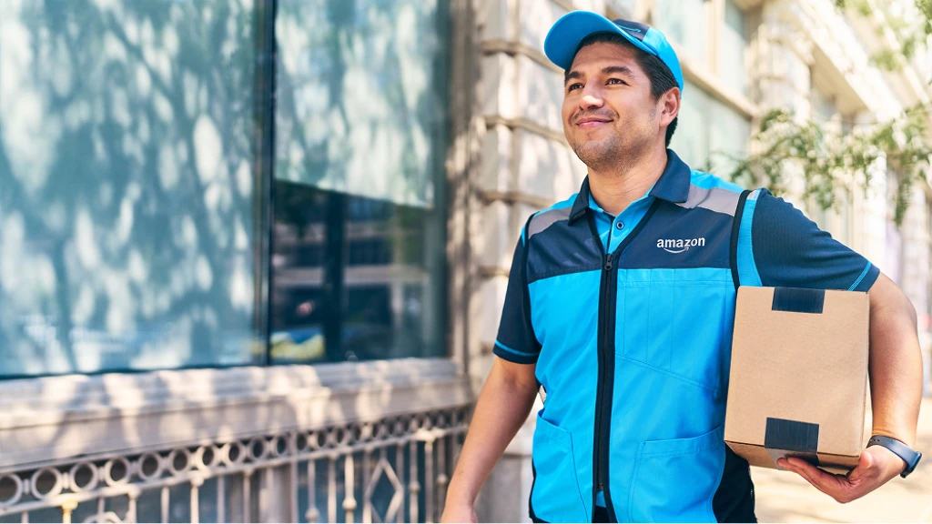 Amazon delivery person in blue uniform carrying a package, standing outside a building in dappled sunlight.